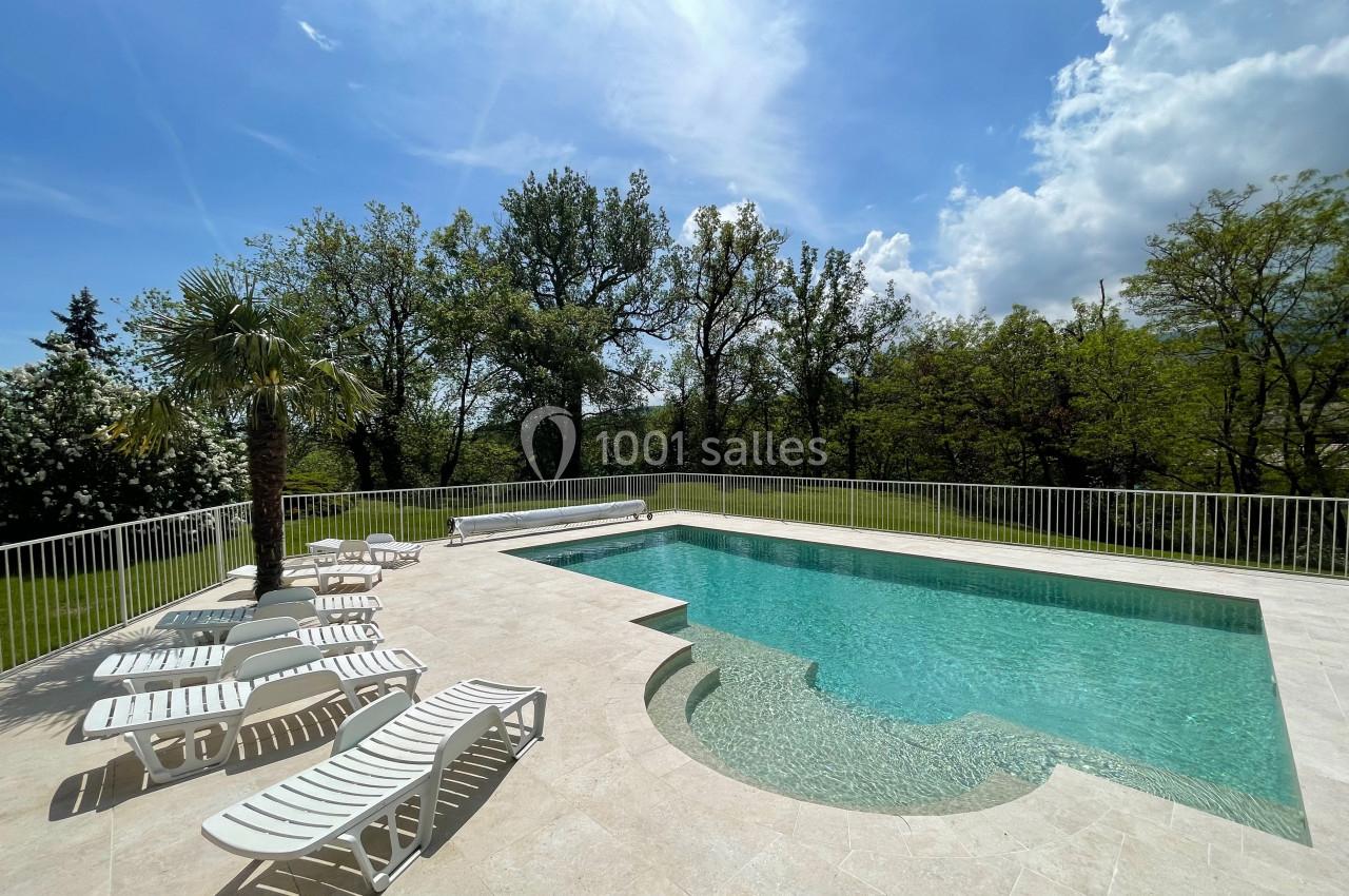Piscine extérieure entourée de chaises longues blanches, avec vue sur des arbres et un ciel partiellement nuageux.
