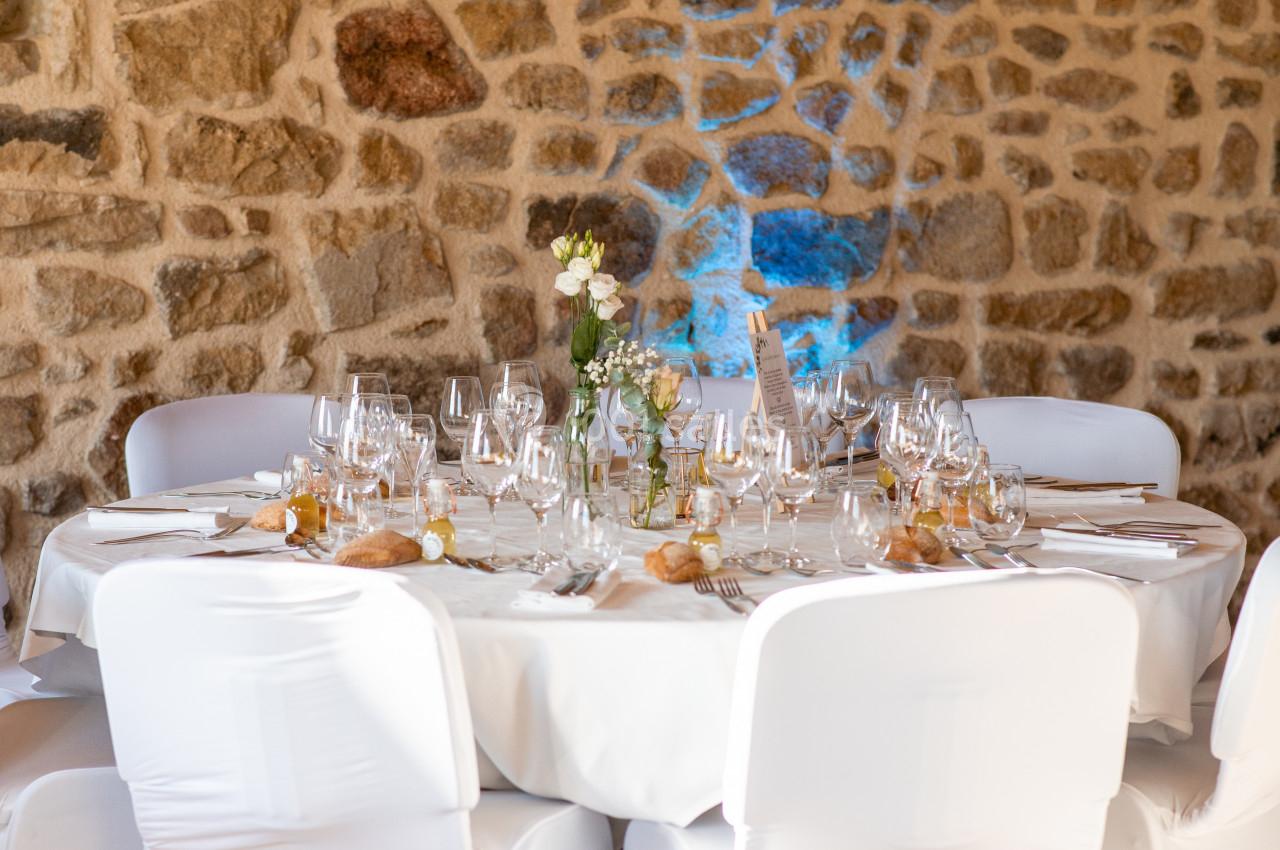 Table ronde élégamment dressée avec nappes blanches, verres, couverts et décoration florale, devant un mur en pierre.