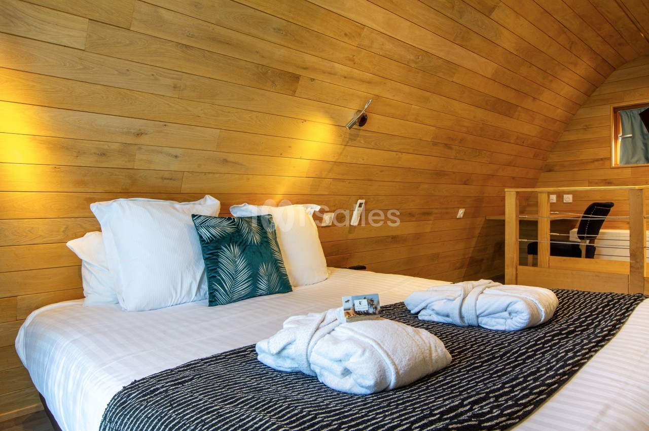 Lit double avec draps blancs, coussins décoratifs, peignoirs pliés et éclairage chaleureux dans une chambre en bois.