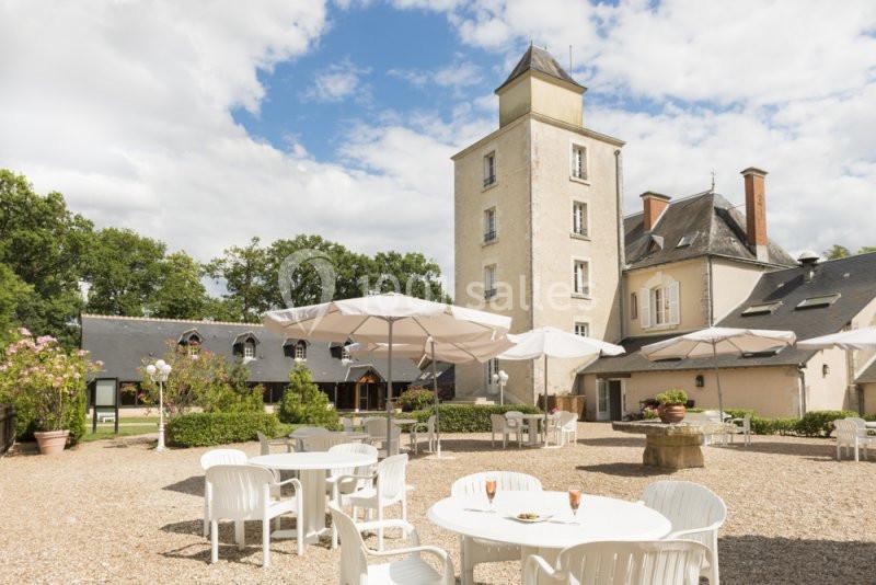 Cour extérieure d'un bâtiment historique avec une tour, des tables blanches, des parasols et un jardin arboré.