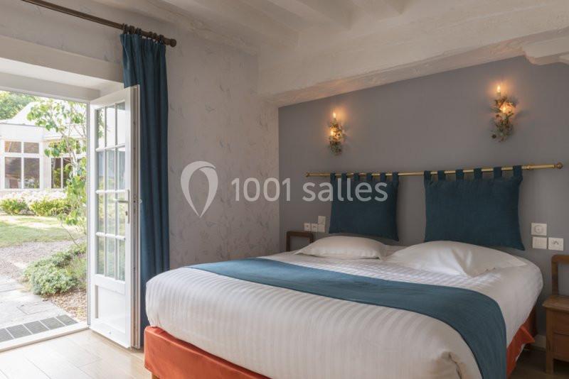 Chambre lumineuse avec lit double, tête de lit bleue, murs gris, deux appliques murales et porte-fenêtre donnant sur un…