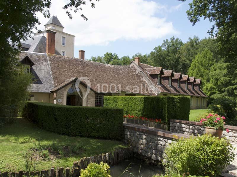 Maison traditionnelle avec toit en tuiles, entourée de haies, d'arbres et d'un petit pont en pierre au premier plan.