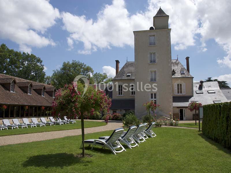 Château avec tour centrale, pelouse aménagée de chaises longues et arbres fleuris sous un ciel partiellement nuageux.