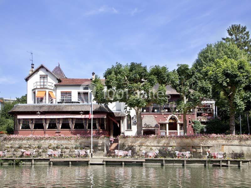Façade d'une maison au bord d'une rivière, entourée d'arbres et de terrasses aménagées avec des tables et des chaises.