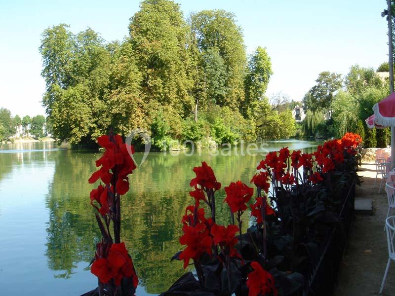 Vue d'une rivière bordée d'arbres avec des fleurs rouges au premier plan et des chaises en terrasse sur la droite.