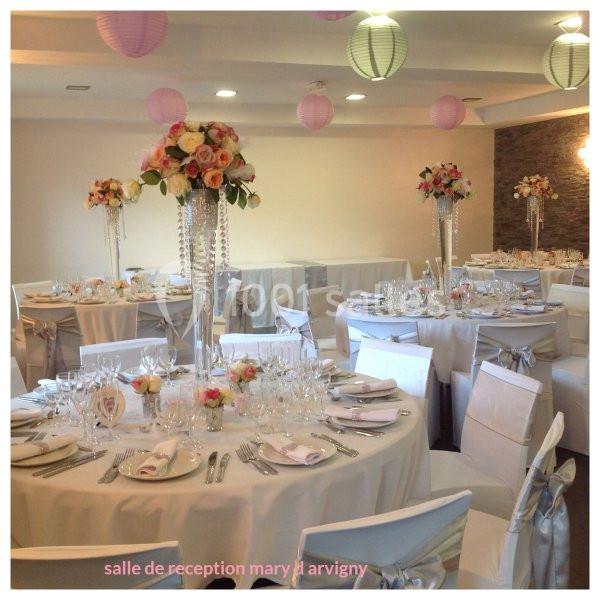 Salle de réception décorée avec des tables rondes, nappes blanches, centres de table fleuris et lanternes suspendues.