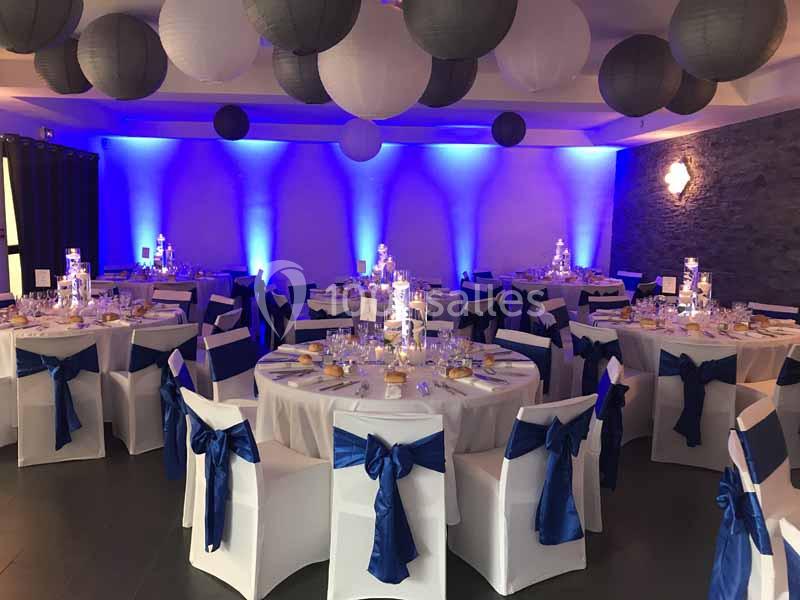 Salle de réception décorée avec des tables rondes, nappes blanches, chaises ornées de nœuds bleus et éclairage tamisé.