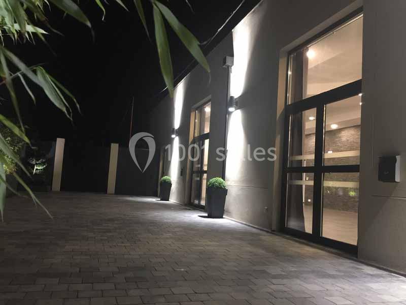 Façade moderne éclairée la nuit, avec de grandes baies vitrées et des plantes en pots sur une allée pavée.