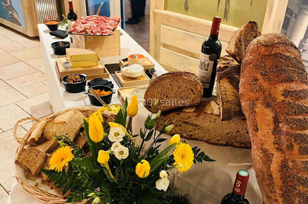 Table garnie de pains variés, charcuteries, fromages, fleurs jaunes et blanches, accompagnée de bouteilles de vin rouge.