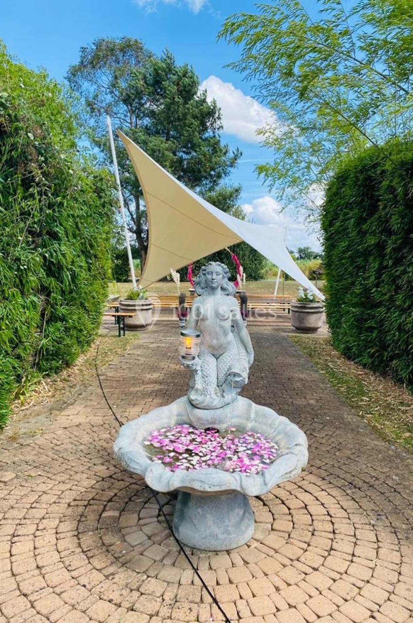 Statue de sirène tenant une lampe, placée dans une fontaine ornée de fleurs, sur une allée pavée entourée de verdure.
