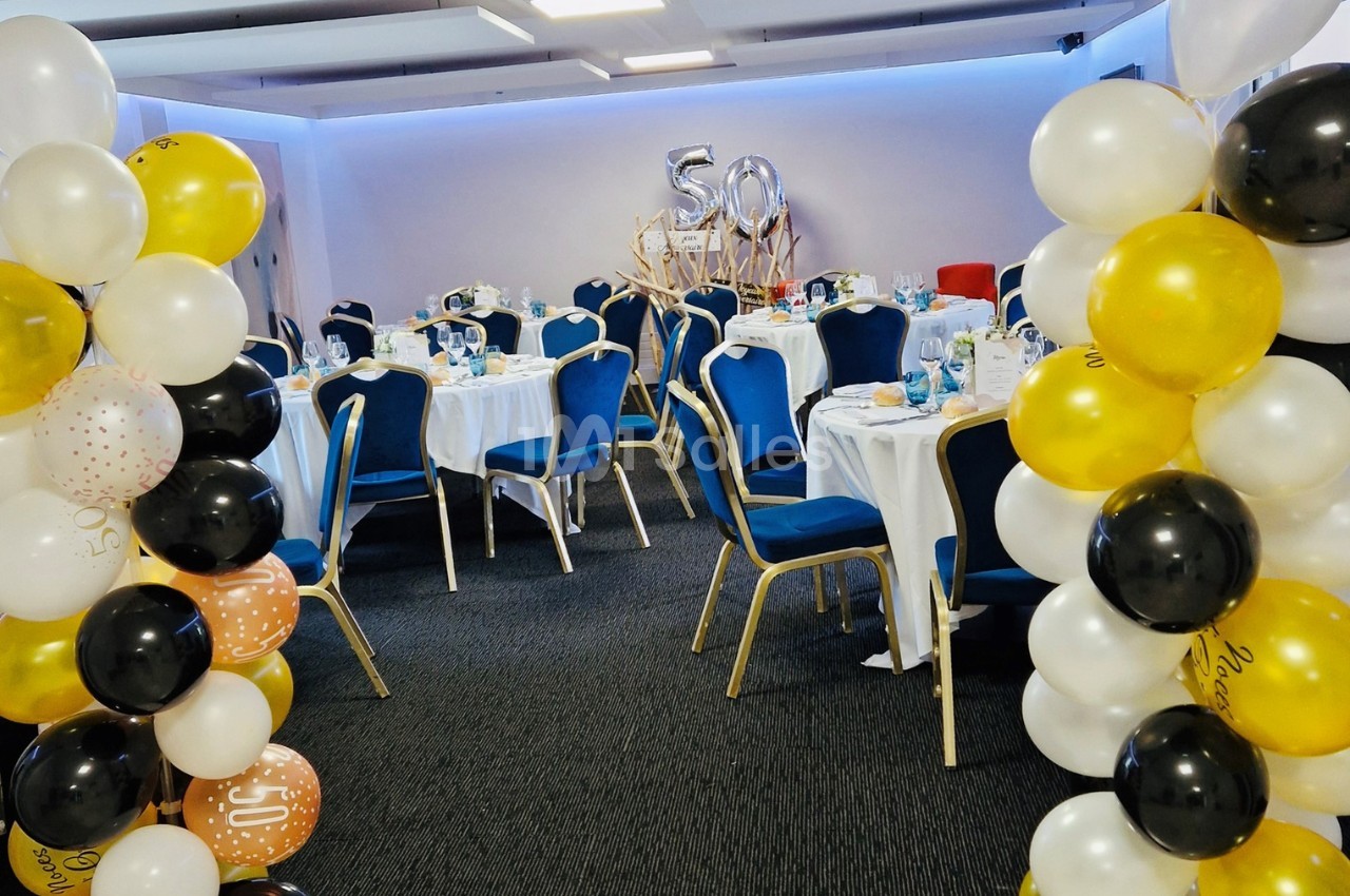 Salle décorée pour une fête avec des tables dressées, des ballons dorés, blancs et noirs, et un chiffre ’50’ en arrière-plan.