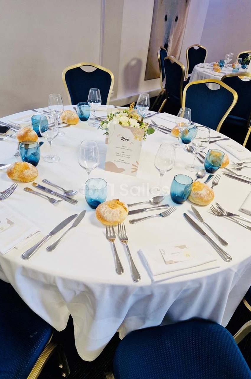 Table ronde dressée avec une nappe blanche, verres bleus, couverts, petits pains et une décoration florale au centre.