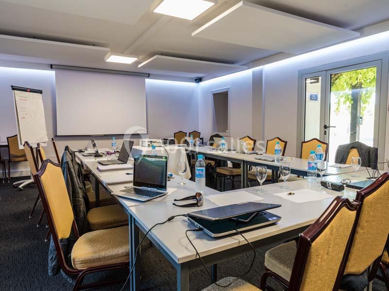 Salle de réunion équipée avec tables en U, chaises, projecteur, ordinateurs portables et bouteilles d'eau.