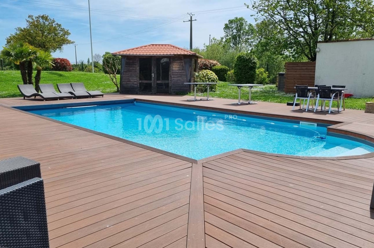 Piscine extérieure entourée d'une terrasse en bois avec chaises longues, mobilier de jardin et cabanon en arrière-plan.