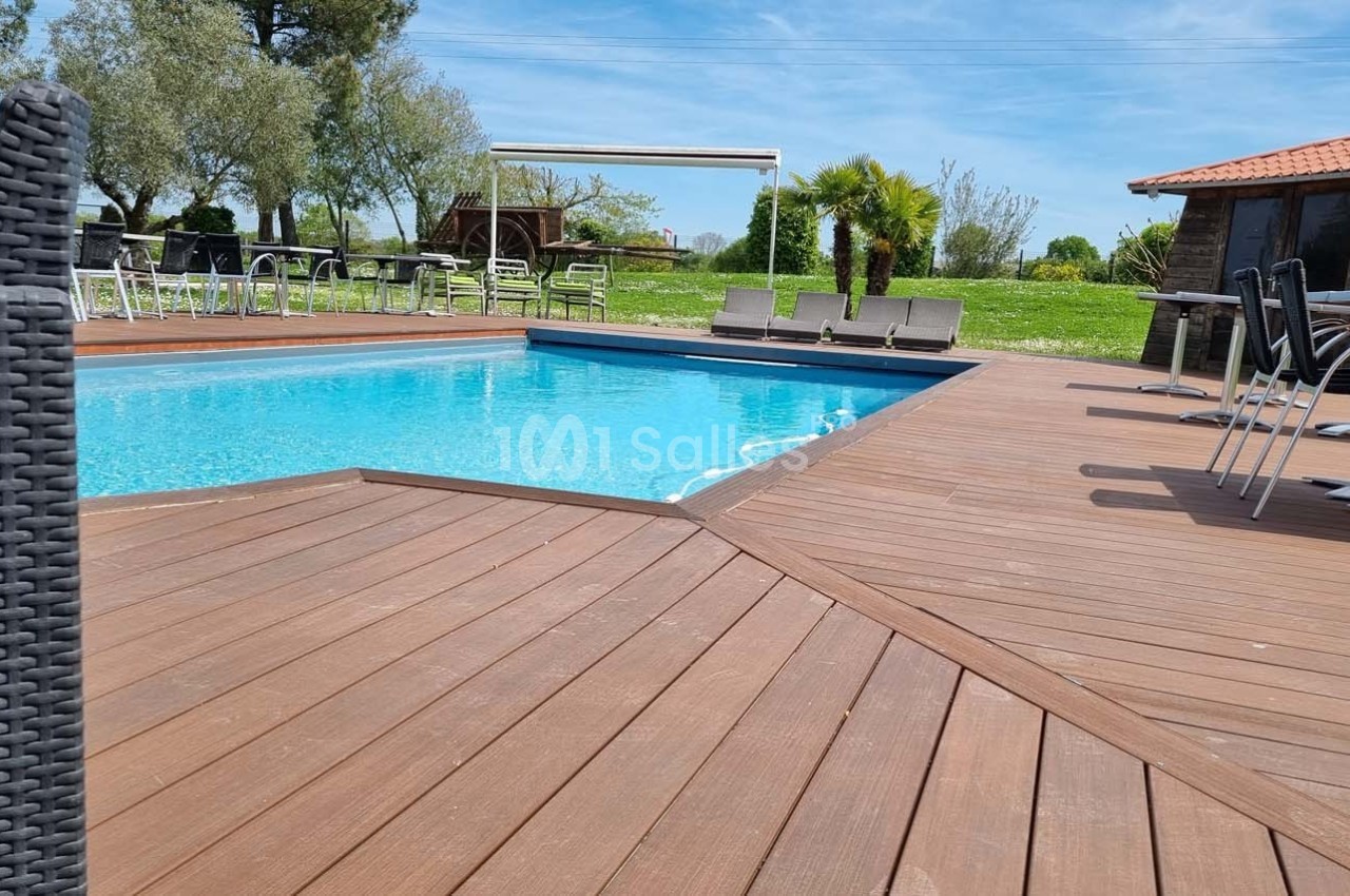 Piscine extérieure entourée d'une terrasse en bois, avec des chaises et des tables, dans un cadre verdoyant.