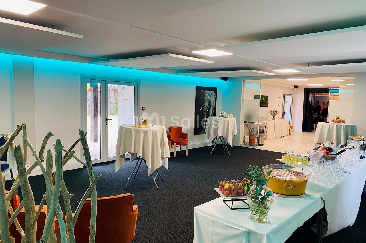 Salle lumineuse aménagée pour un événement, avec tables hautes, buffet et décoration moderne.