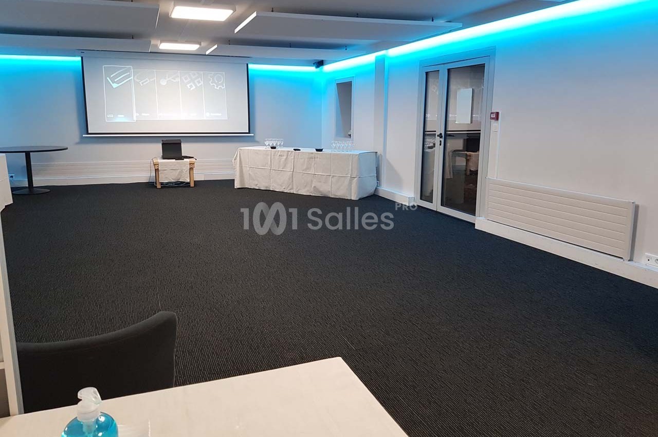 Salle de réunion vide avec un projecteur allumé, des tables couvertes de nappes blanches et un distributeur de gel visible.