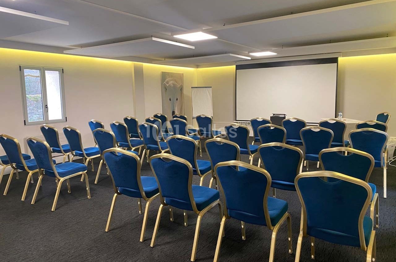 Salle de conférence avec des rangées de chaises bleues face à un écran de projection et un tableau blanc.