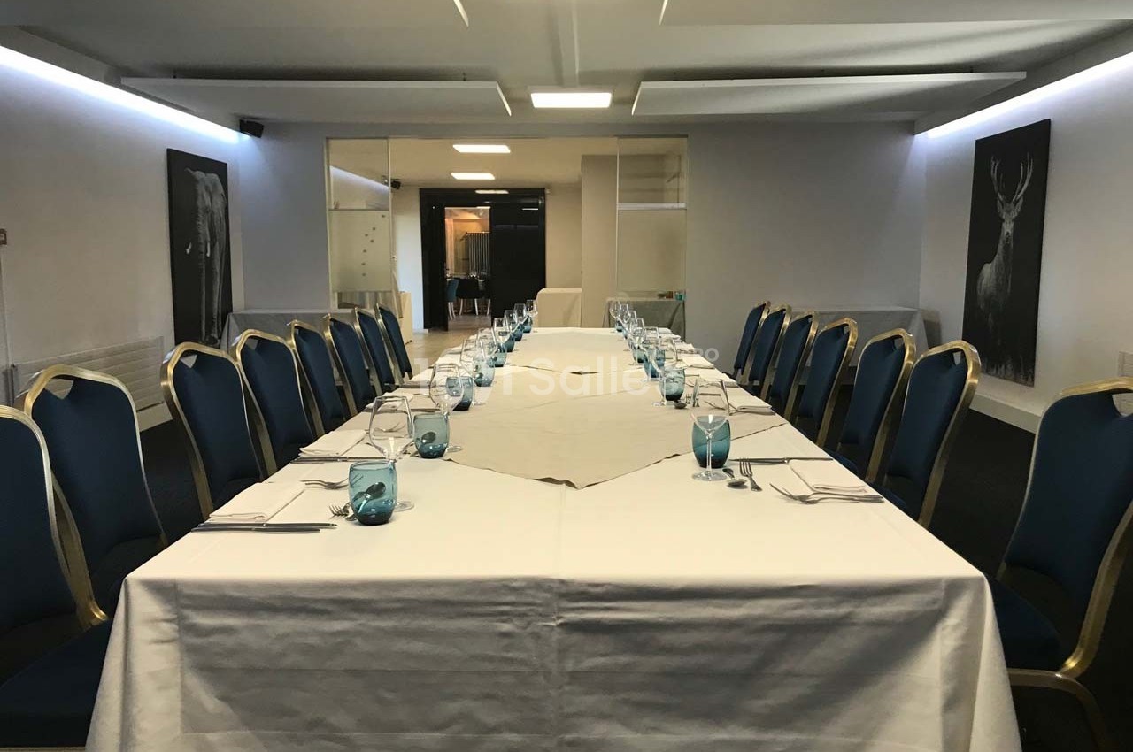 Salle de réunion avec une grande table en U, nappée de blanc, entourée de chaises bleues et décorée de verres turquoise.