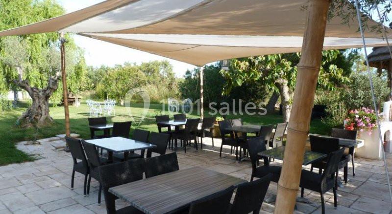 Terrasse extérieure avec tables et chaises sous des voiles d'ombrage, entourée de verdure et d'arbres.