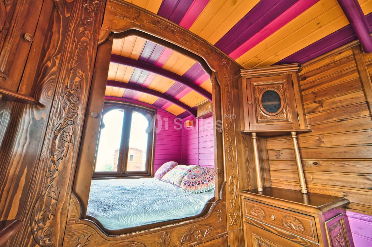 Intérieur d'une roulotte en bois avec un lit coloré, des motifs sculptés et un plafond aux teintes vives.