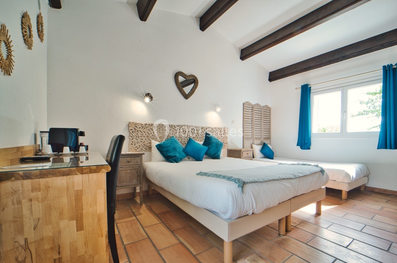 Chambre lumineuse avec deux lits, décorée de coussins bleus, poutres apparentes et bureau en bois.