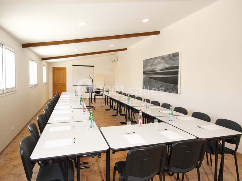 Salle de réunion lumineuse avec tables disposées en U, chaises noires, tableau blanc et bouteille d'eau sur les tables.