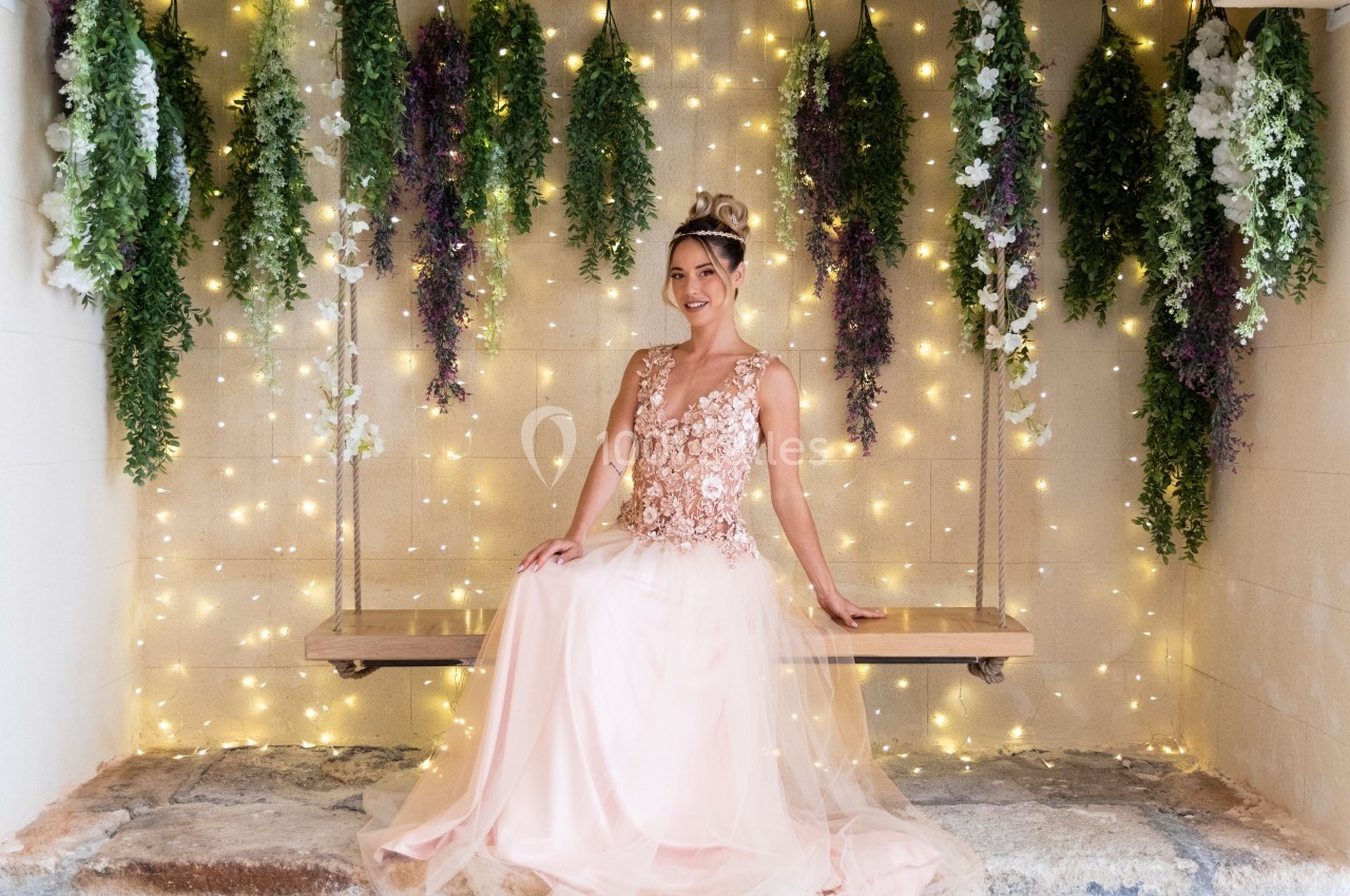 Femme en robe rose ornée de fleurs assise sur une balançoire, entourée de guirlandes lumineuses et de plantes suspendues.