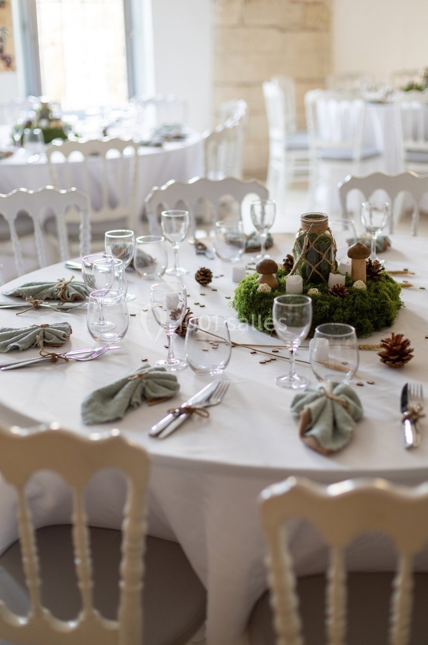 Table ronde décorée pour un événement, avec nappes blanches, serviettes pliées, verres, couverts et centre de table végétal.