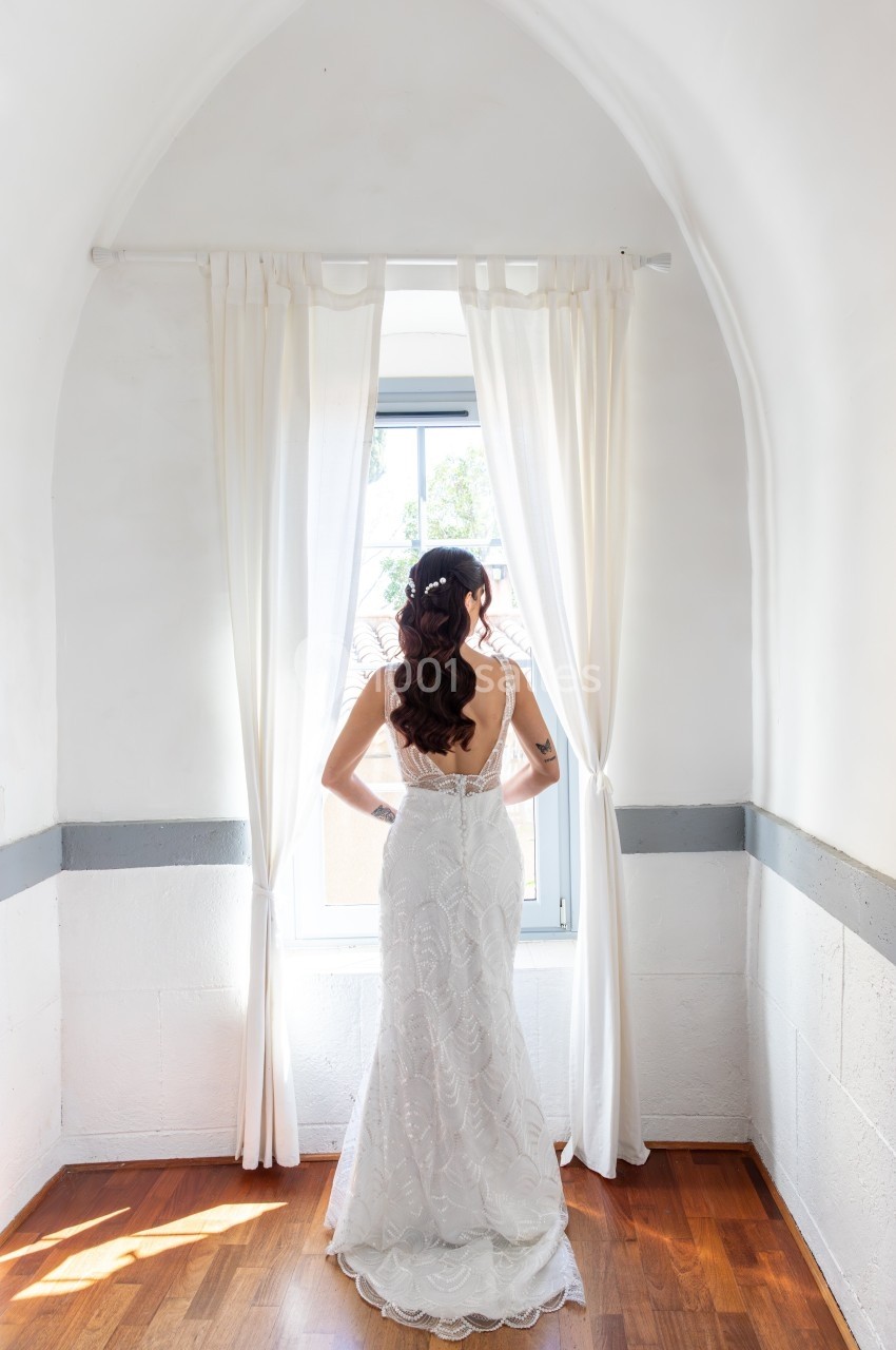 Femme en robe de mariée blanche vue de dos, regardant par une fenêtre encadrée de rideaux blancs dans une pièce lumineuse.