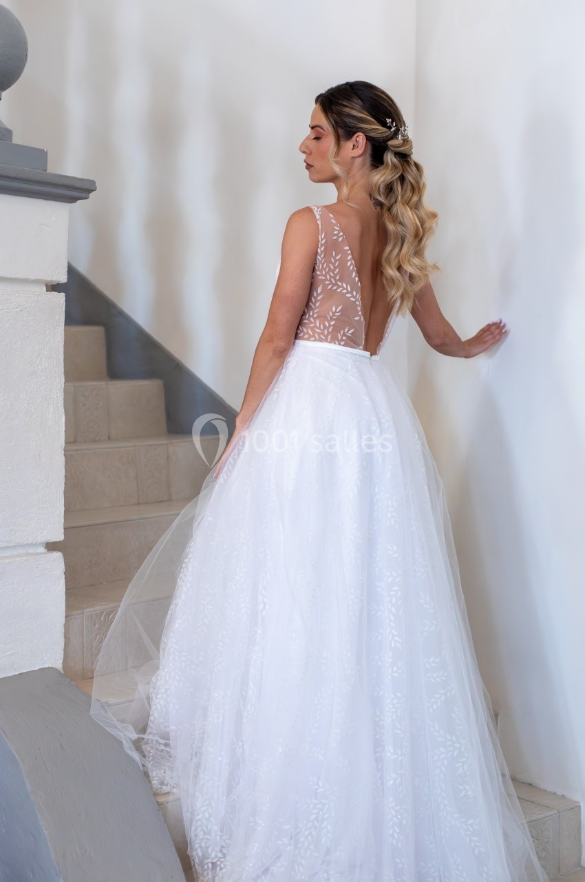 Femme en robe de mariée blanche avec dos en dentelle transparente, montant un escalier intérieur en pierre.