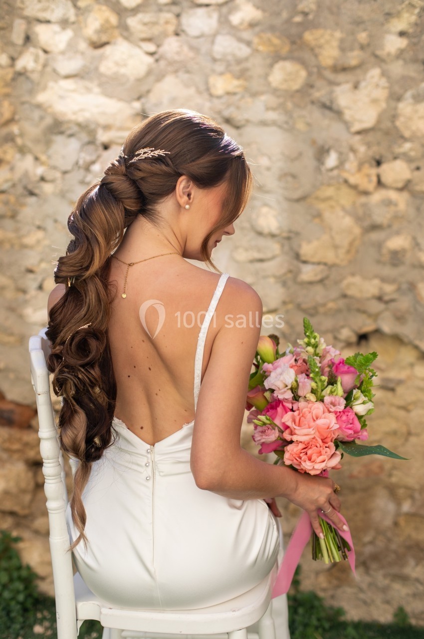 Femme en robe de mariée blanche, de dos, tenant un bouquet de fleurs colorées devant un mur en pierre.