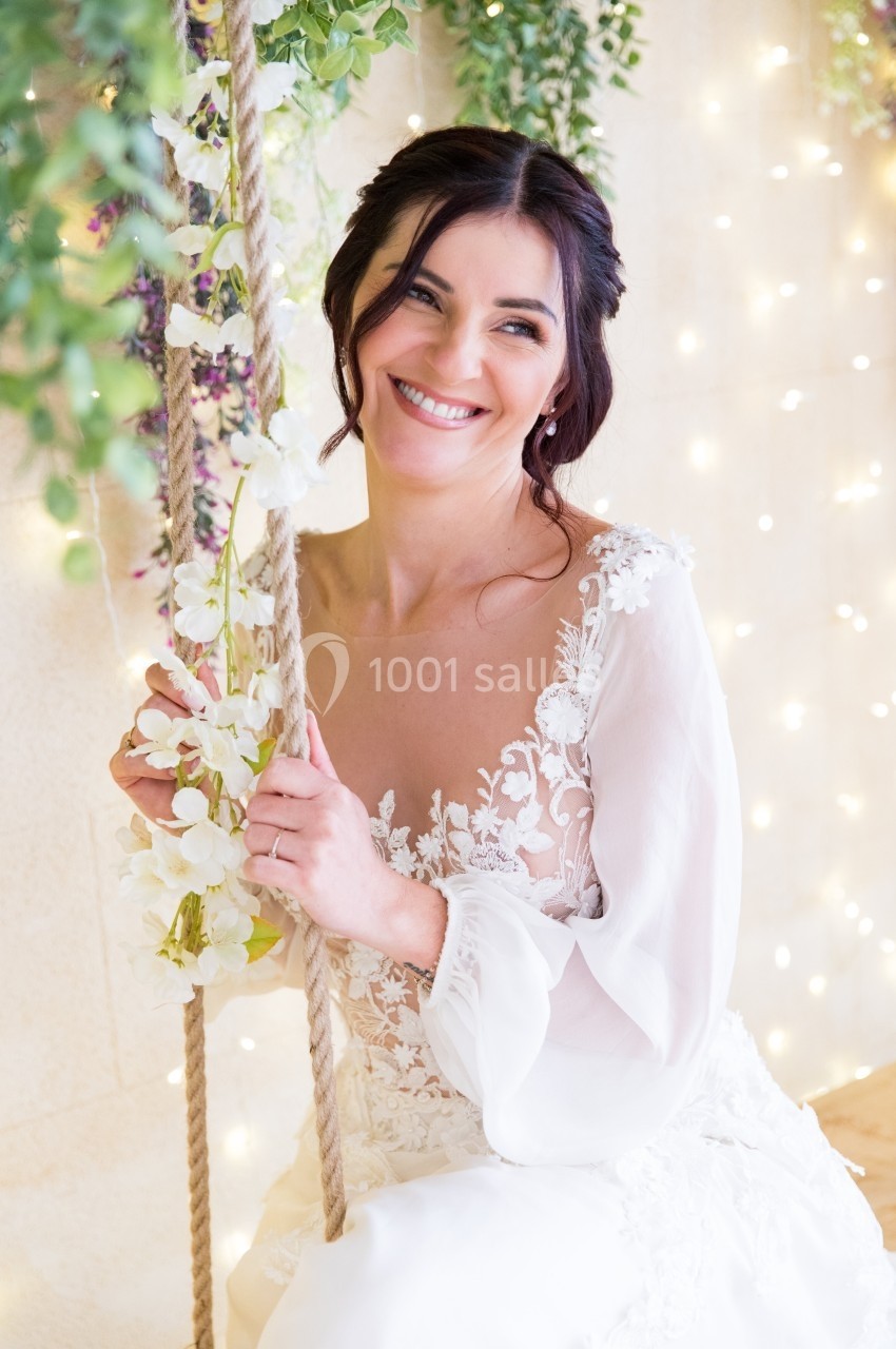 Une femme souriante en robe blanche ornée de dentelle est assise sur une balançoire décorée de fleurs et de feuillage.