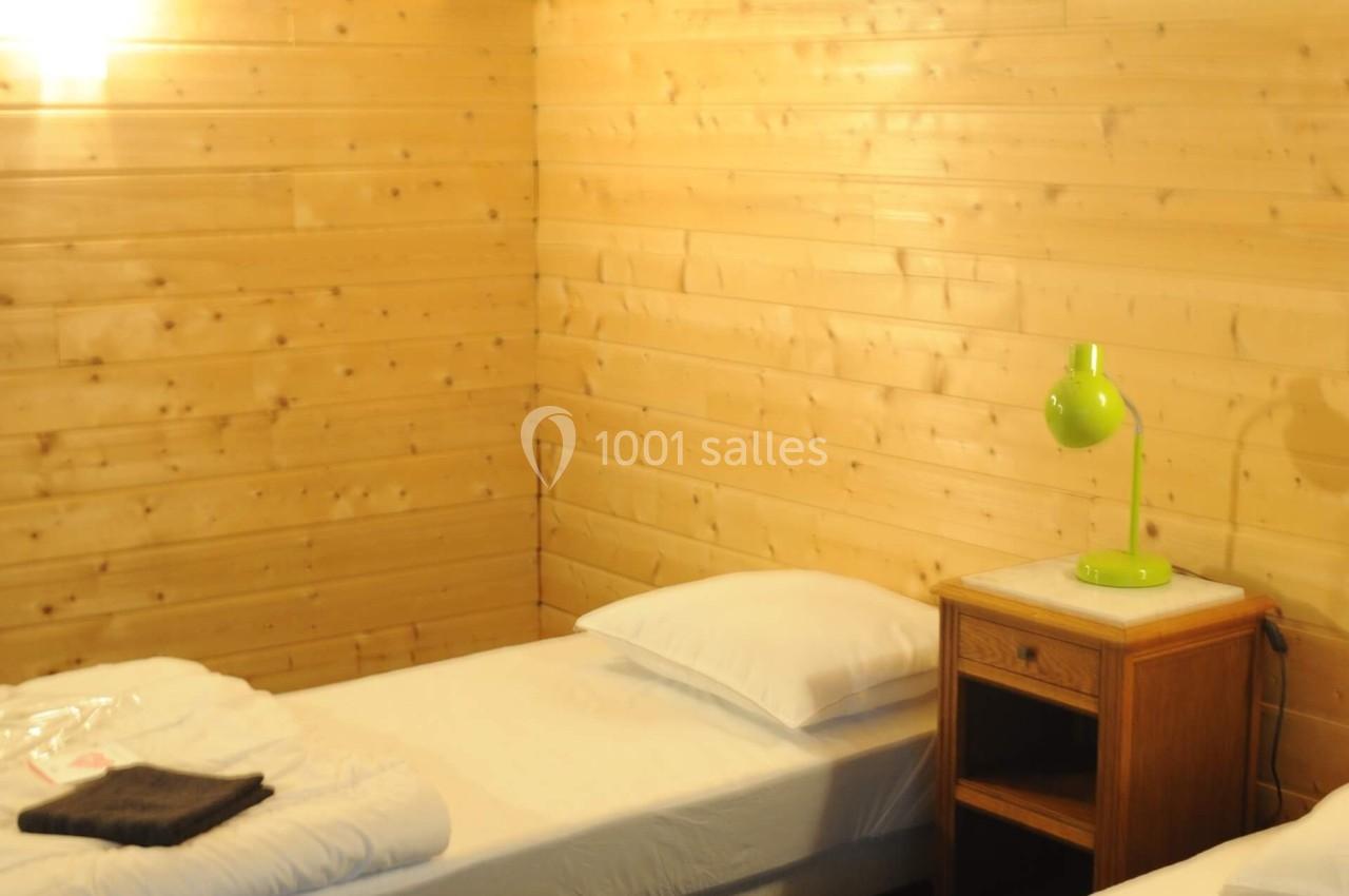 Deux lits simples avec draps blancs dans une chambre en bois clair, éclairée par une lampe murale et une lampe de chevet…