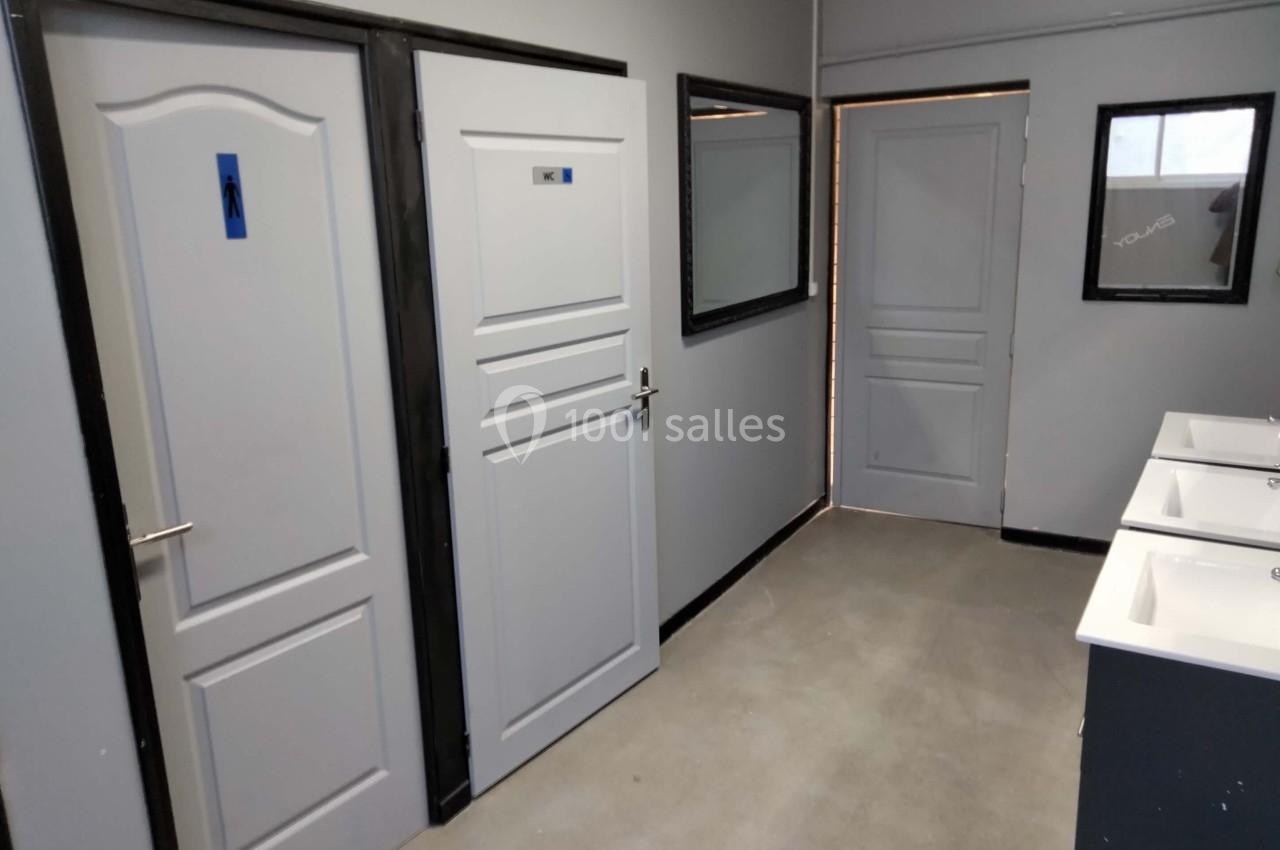 Vue d'un espace sanitaire avec portes de toilettes, lavabos alignés et miroirs sur les murs gris.