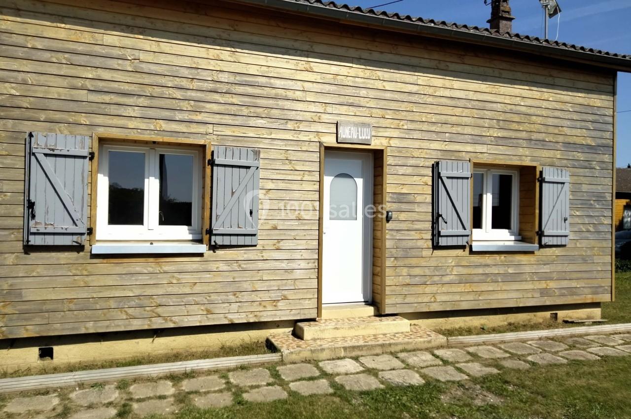 Façade d'une maison en bois avec trois fenêtres à volets gris et une porte blanche, entourée d'une pelouse.