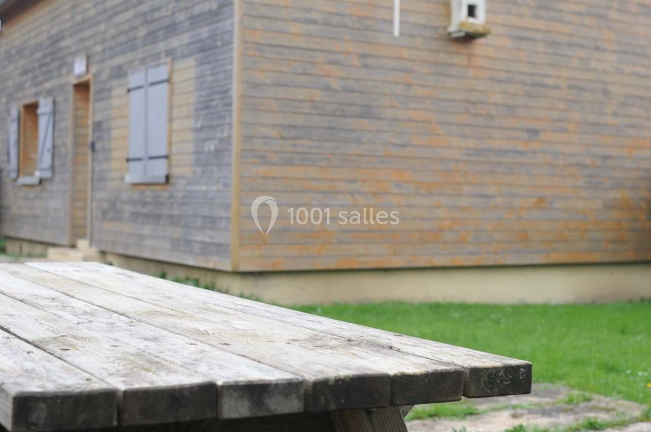 Table de pique-nique en bois devant une maison en bois avec des volets fermés, entourée de pelouse.