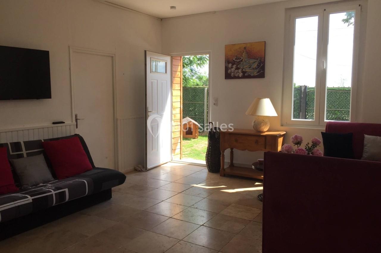 Salon lumineux avec canapé, table, lampe, tableau au mur, porte ouverte sur un jardin et fenêtre laissant entrer la lumière.