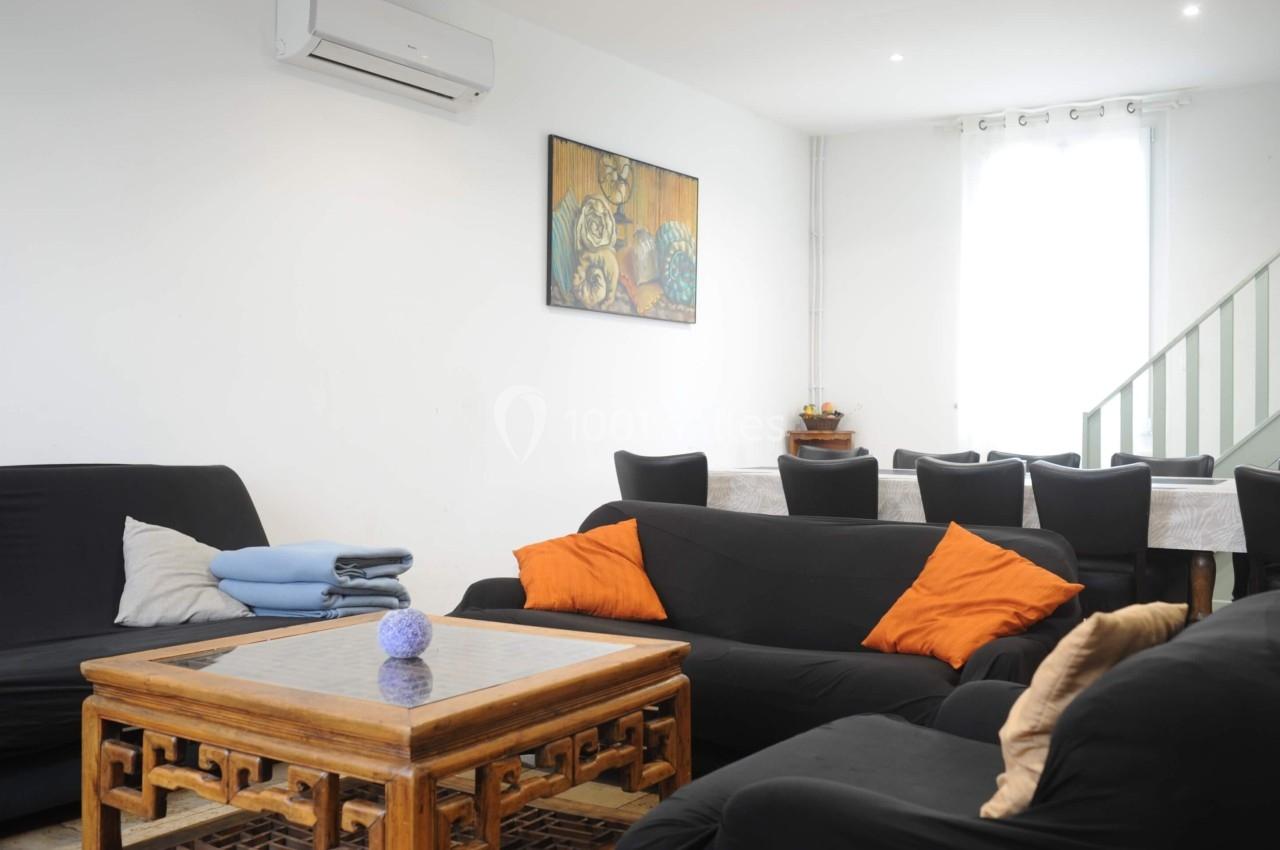 Salon lumineux avec canapés noirs, coussins colorés, table basse en bois, tableau mural et salle à manger en arrière-plan.