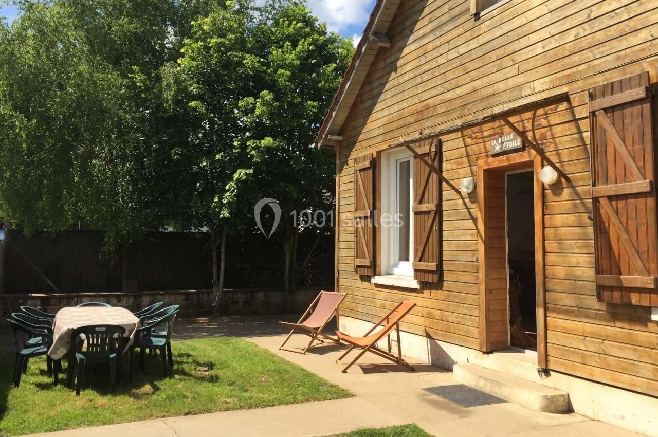 Maison en bois avec volets ouverts, entourée de verdure, table et chaises de jardin sur une pelouse ensoleillée.
