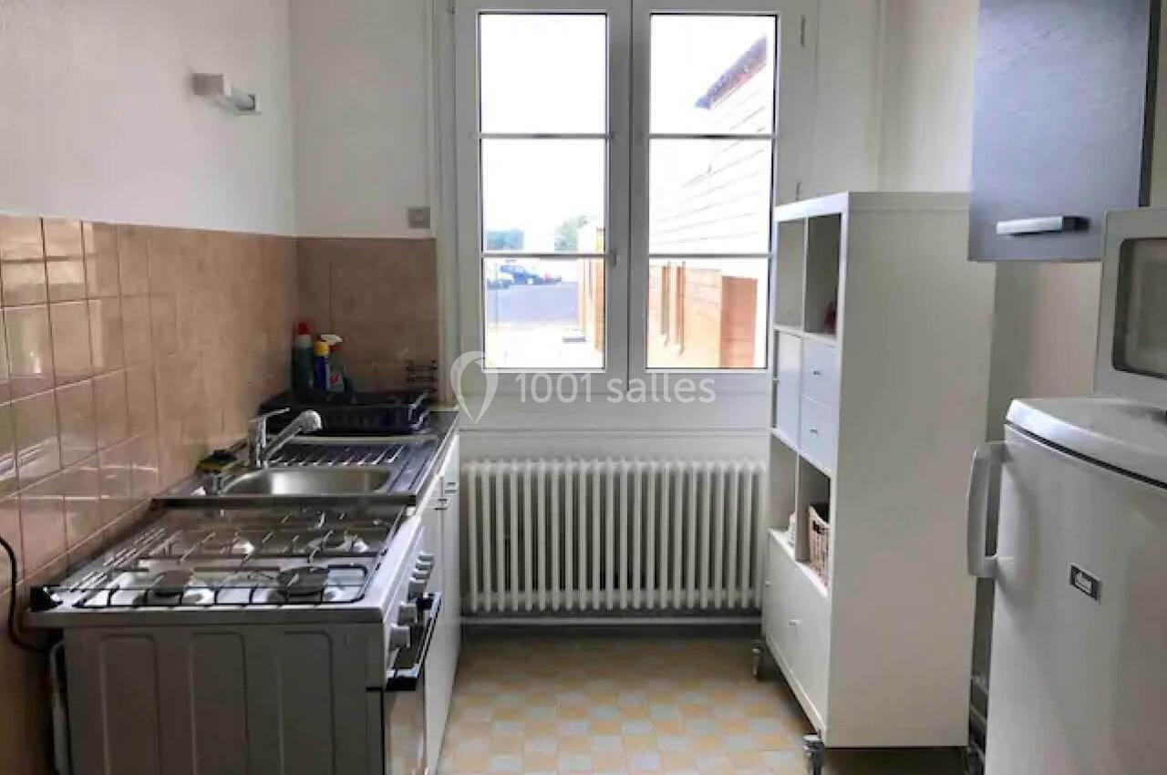 Cuisine lumineuse avec évier, cuisinière, réfrigérateur, étagère blanche et fenêtre donnant sur l'extérieur.