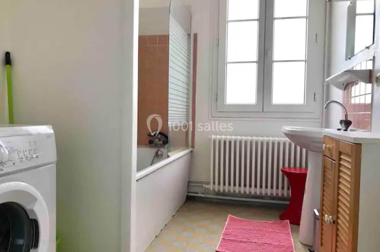 Salle de bain lumineuse avec baignoire, lavabo, machine à laver, tapis rose et fenêtre à double battant.