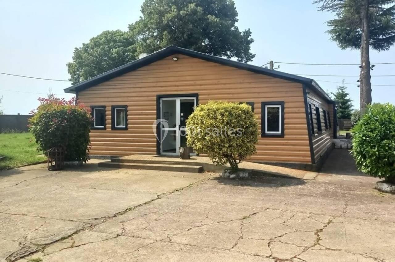 Bâtiment en bois avec plusieurs fenêtres, entouré de végétation, situé sur une surface bétonnée.