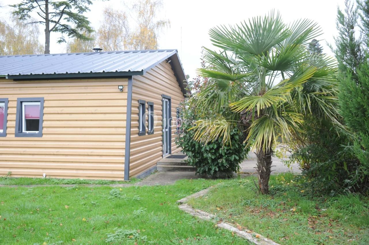 Façade latérale d'une maison en bois avec un toit métallique, entourée de végétation et d'un palmier au premier plan.