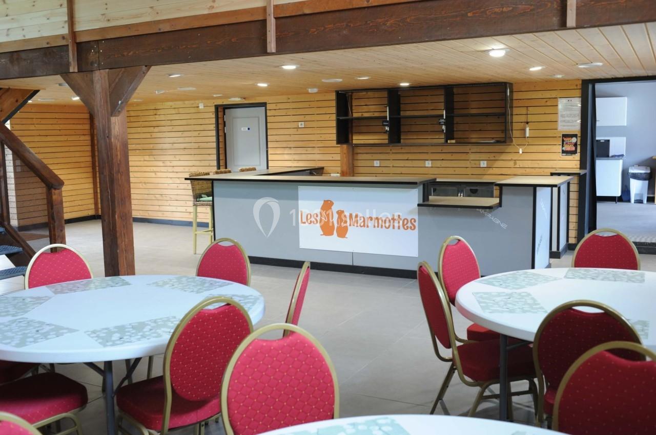 Salle avec des tables rondes, chaises rouges et un comptoir de bar en bois portant le logo ’Les Marmottes’.
