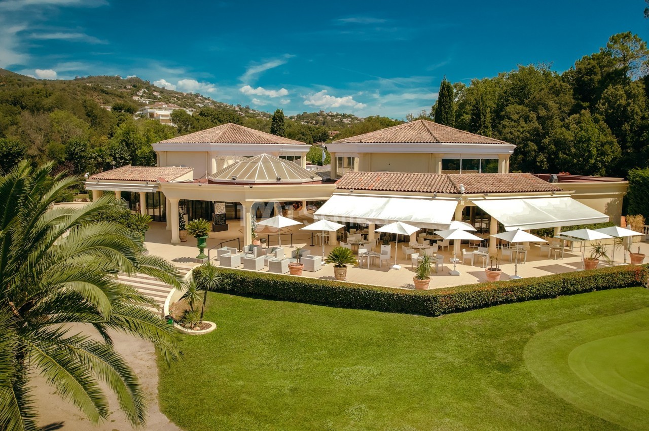 Vue aérienne d'un bâtiment lumineux entouré de verdure, avec une terrasse aménagée et des parasols blancs.
