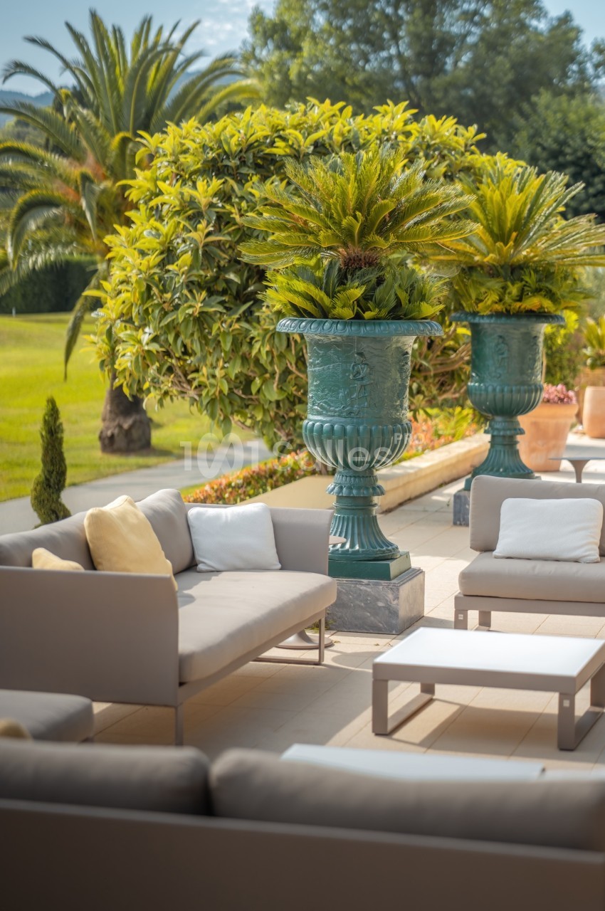 Mobilier de jardin moderne avec canapés gris et tables basses, entouré de plantes et de grands vases décoratifs.