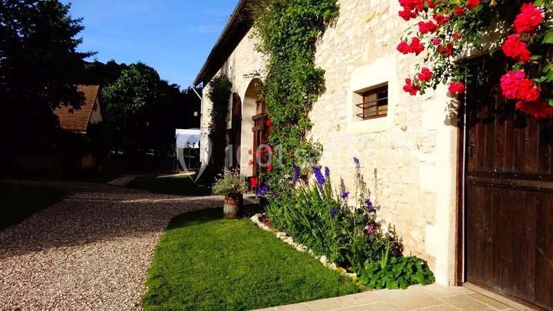 Façade en pierre d'une maison avec fleurs colorées, allée en gravier et pelouse sous un ciel dégagé.