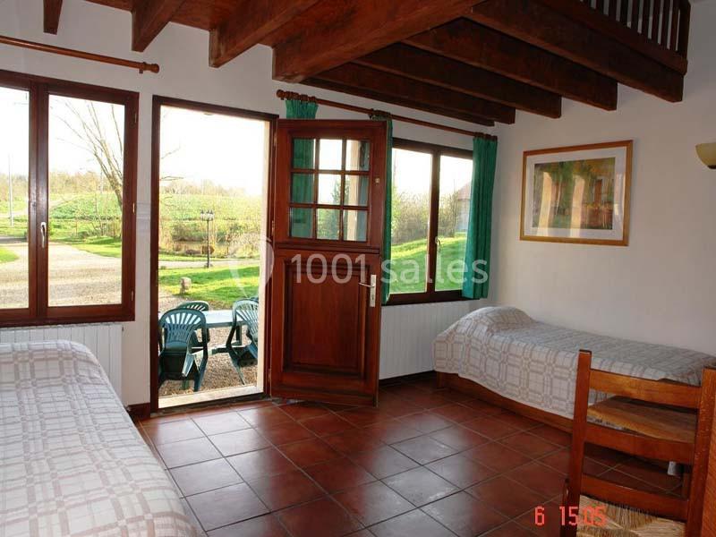 Chambre lumineuse avec deux lits simples, sol en carrelage, plafond en bois, et vue sur un jardin depuis une porte ouverte.