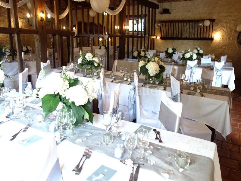 Salle de réception décorée avec des tables dressées, nappes blanches, fleurs et chaises couvertes.